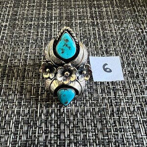 May include: A silver ring featuring two teardrop-shaped turquoise stones with dark matrix patterns, accented by three metal flowers and leaf-like engravings. A small white square tag with the handwritten number 6 is placed next to the ring.