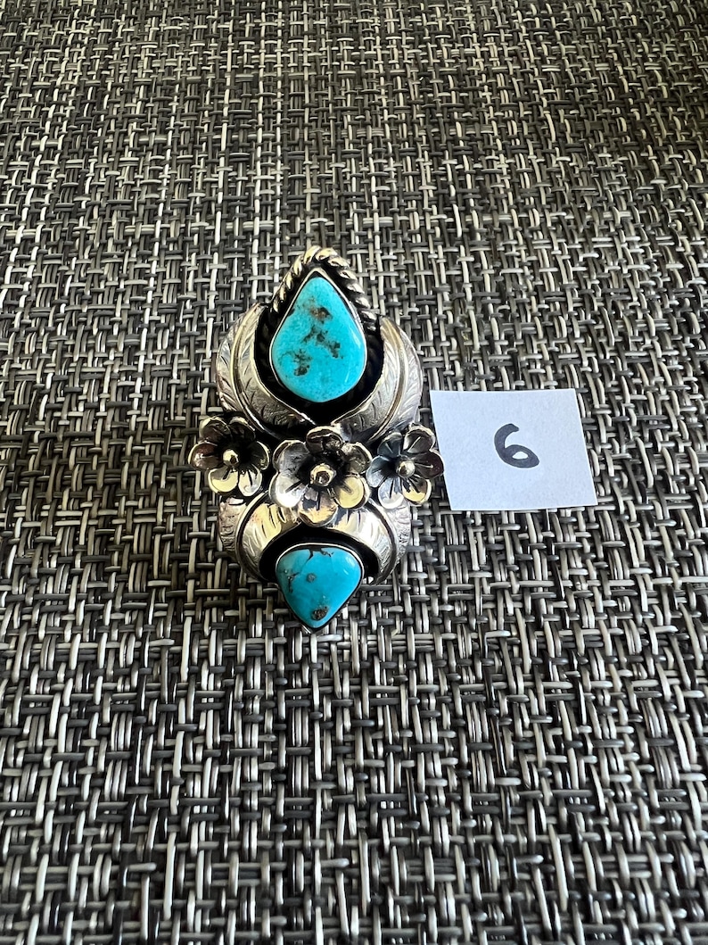 May include: A silver ring featuring two teardrop-shaped turquoise stones with dark matrix patterns, accented by three metal flowers and leaf-like engravings. A small white square tag with the handwritten number 6 is placed next to the ring.
