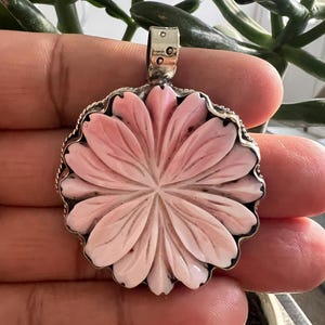 Puede incluir: Un colgante circular que presenta una flor tallada de color rosa claro con múltiples pétalos, engastada en un marco de metal plateado con un borde de cuerda trenzada y una pequeña anilla en la parte superior para colgar de una cadena.