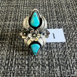 May include: A silver ring featuring two teardrop-shaped turquoise stones, one at the top and one at the bottom, separated by three metal flowers in the center. The metal surfaces are engraved with leaf patterns and a small white tag with the number 1.