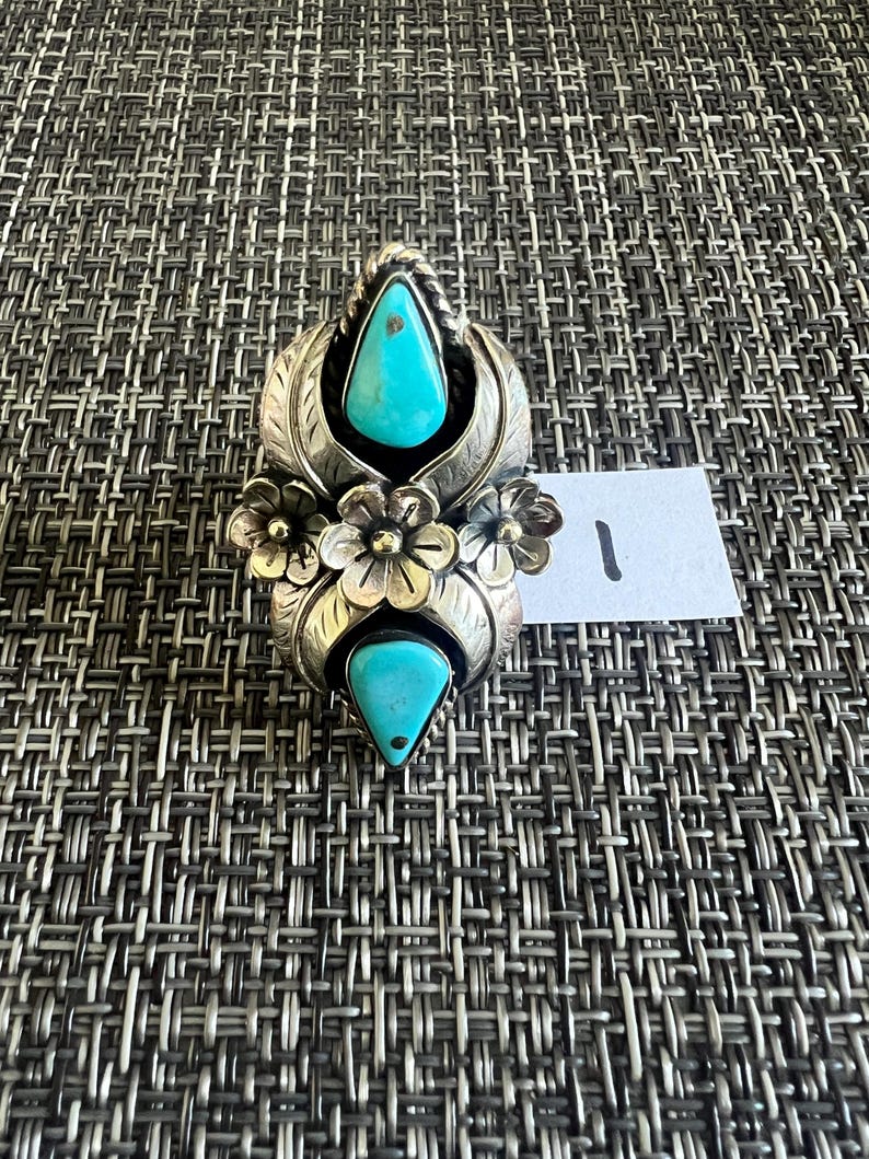 May include: A silver ring featuring two teardrop-shaped turquoise stones, one at the top and one at the bottom, separated by three metal flowers in the center. The metal surfaces are engraved with leaf patterns and a small white tag with the number 1.