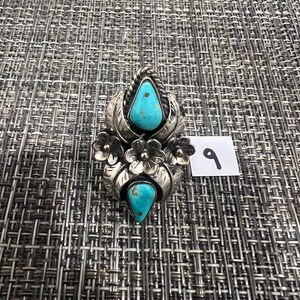 May include: A silver ring featuring two teardrop-shaped turquoise stones, decorative metal leaves, and small flower accents. The ring is positioned next to a small white square tag with the handwritten number 9 in black ink.