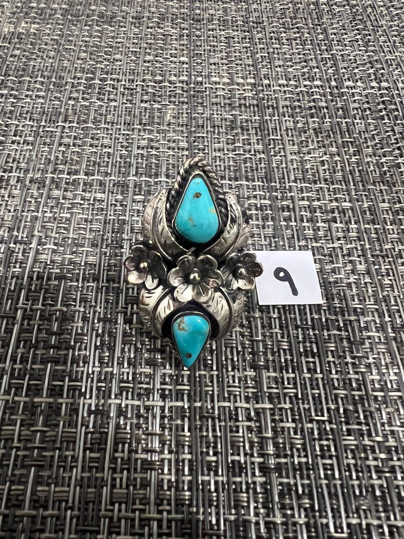 May include: A silver ring featuring two teardrop-shaped turquoise stones, decorative metal leaves, and small flower accents. The ring is positioned next to a small white square tag with the handwritten number 9 in black ink.