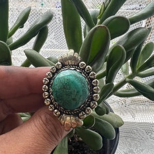 May include: A silver ring with a large, round, turquoise stone. The ring has a detailed, ornate setting with a scalloped top and small decorative elements around the stone. The ring is held in front of a background of green plants.