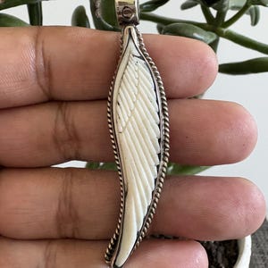 May include: A long, slender pendant shaped like a bird wing, featuring a carved off-white material inlay set within a polished silver frame. The edge of the frame has a twisted rope texture, and a silver bail is attached to the top for hanging.