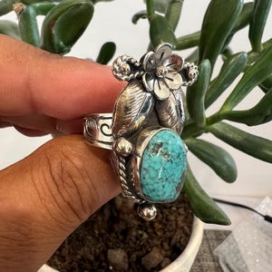 May include: A sterling silver ring featuring an oval turquoise stone with dark matrix patterns. The ring is decorated with a metal flower and leaf motif at the top, along with twisted wire detailing and engraved scrollwork on the band.