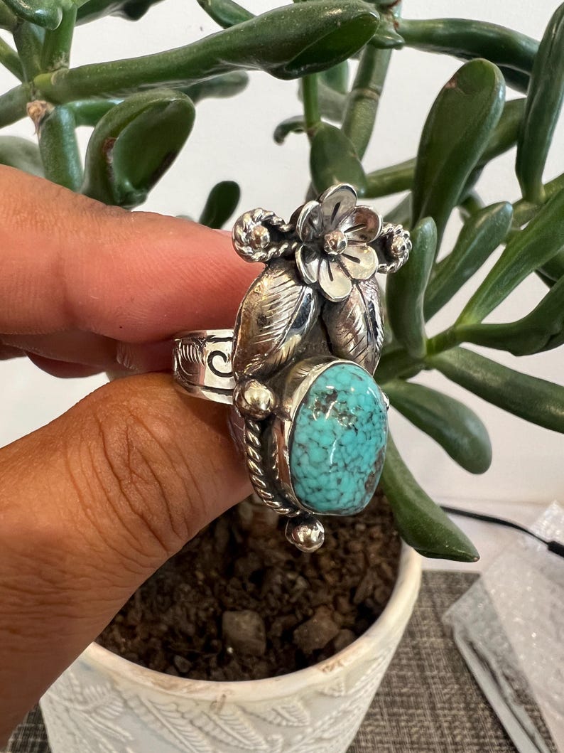 May include: A sterling silver ring featuring an oval turquoise stone with dark matrix patterns. The ring is decorated with a metal flower and leaf motif at the top, along with twisted wire detailing and engraved scrollwork on the band.