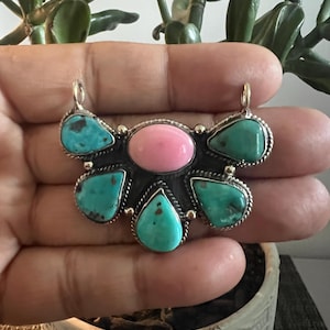 May include: A pendant featuring a central oval pink stone surrounded by five teardrop-shaped turquoise stones with dark matrix inclusions. The stones are set in a silver frame with twisted wire detailing and two top loops for attachment to a chain.