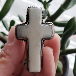Op de afbeelding: Een witte kruisvormige hanger met een zilverkleurige, touwachtige rand. Het kruis wordt in een hand gehouden, met een groene plant op de achtergrond. Het kruis heeft een glad, gebroken witte oppervlak.