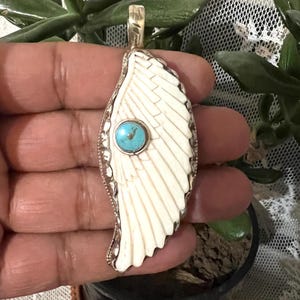 May include: A pendant with an ivory-coloured wing design, a silver-tone border, and a turquoise stone. The wing is carved. The pendant is held in a hand, with a gold-tone bail.