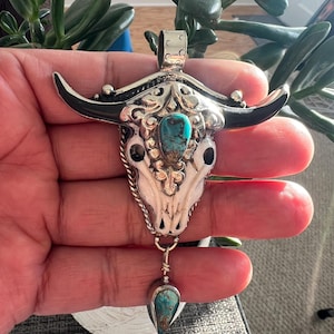 May include: A silver pendant in the shape of a bull skull, featuring black horns and a turquoise stone in the center. The pendant has intricate detailing and a teardrop-shaped turquoise stone hanging below. The pendant is held in a hand.