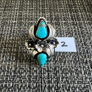 May include: A silver ring featuring two teardrop-shaped turquoise stones, one at the top and one at the bottom, accented by three central silver flowers and leaf motifs. A small white paper tag with the handwritten number 2 is placed beside it.