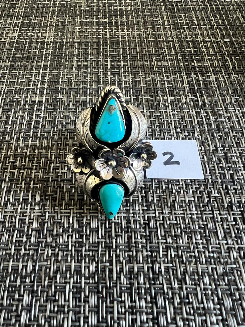 May include: A silver ring featuring two teardrop-shaped turquoise stones, one at the top and one at the bottom, accented by three central silver flowers and leaf motifs. A small white paper tag with the handwritten number 2 is placed beside it.