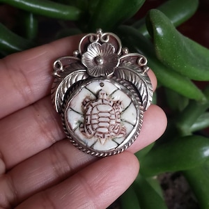 May include: A silver pendant featuring a carved turtle design on a white background. The pendant is surrounded by a silver frame with a floral design.