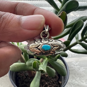 May include: A silver pendant with a turquoise stone center. The pendant has an ornate design with leaf-like details. The pendant is held in front of a potted plant with green leaves.