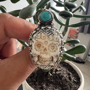 May include: A silver pendant with a carved skull design, heart-shaped eye sockets, and a turquoise stone. The pendant is held in front of a green plant in a white pot.