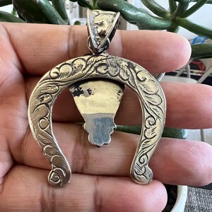 May include: A silver-toned metal pendant shaped like an inverted horseshoe with intricate engraved scrollwork patterns along the curved frame. The pendant features a central triangular element with a dark, smooth inlay and a large, textured bail at the top.