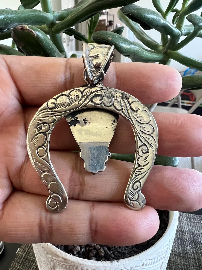 May include: A silver-toned metal pendant shaped like an inverted horseshoe with intricate engraved scrollwork patterns along the curved frame. The pendant features a central triangular element with a dark, smooth inlay and a large, textured bail at the top.