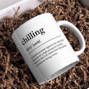 Könnte beinhalten: Eine weiße Keramiktasse mit einer schwarzen Definition des Wortes "chilling" darauf gedruckt. Die Definition lautet: "Ein friedlicher Protest gegen die unerbittlichen Anforderungen einer hochdruckorientierten Gesellschaft, basierend auf der festen Überzeugung, dass weniger tun manchmal mehr bedeutet."