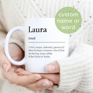 Puede incluir: Una taza de cerámica blanca con texto impreso en negro que define el nombre Laura como un sustantivo con cuatro descripciones numeradas que incluyen amable, talentosa, generosa, hace los mejores rollos de canela, baila, ama el café.