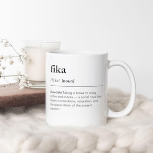 Op de afbeelding: Witte keramische mok met het woord "fika" in het zwart gedrukt, samen met de uitspraak en definitie. Een kaars en decoratieve bloemen staan op de achtergrond. De mok is een sociaal ritueel om van koffie en snacks te genieten.