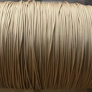 Puede incluir: Un carrete de cordón beige, bien enrollado y cuidadosamente dispuesto. El cordón parece estar hecho de un material liso, posiblemente sintético, y está muy apretado. El color es beige neutro, adecuado para varios proyectos de manualidades.