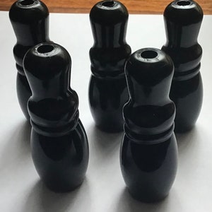 Puede incluir: Cinco bolos negros brillantes dispuestos sobre una superficie blanca. Los bolos tienen una forma clásica con una base redondeada, una sección media con un anillo y una parte superior cilíndrica con un agujero.