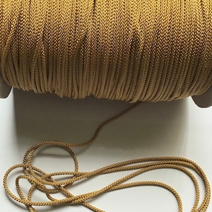 Puede incluir: Una bobina de cordón de color dorado, con un tramo de cordón dispuesto sobre una superficie blanca.