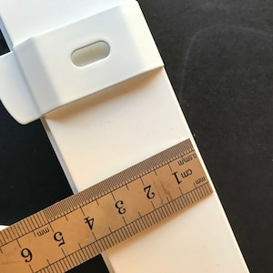 May include: Close-up of a white plastic component with a rectangular shape and a curved edge. A ruler is placed across the component, showing measurements in centimetres. The component appears to be part of a larger structure.