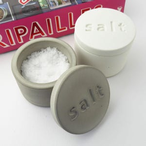 May include: A grey concrete salt cellar with a matching lid, next to a white salt container. The open cellar is filled with coarse white salt. The word "salt" is embossed on the lid and the white container.