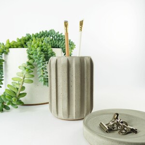 May include: A grey, ribbed concrete pencil holder with a pencil and a paintbrush inside. A small, round concrete dish with silver binder clips sits in front of the holder. A white planter with a trailing green succulent is in the background.