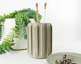 May include: A gray, ribbed concrete pencil holder with a pencil and a paintbrush inside. A small, round concrete dish with silver binder clips sits in front of the holder. A white planter with a trailing green succulent is in the background.