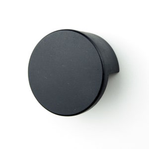 May include: Black round cabinet knob with a slightly curved edge. The knob is made of metal and has a matte finish.