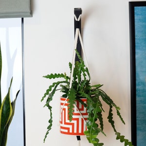 Puede incluir: Un macetero colgante con un cord&oacute;n de macram&eacute; blanco y un gancho de metal negro. El macetero es naranja y blanco con un patr&oacute;n geom&eacute;trico. Una planta verde est&aacute; creciendo en el macetero.