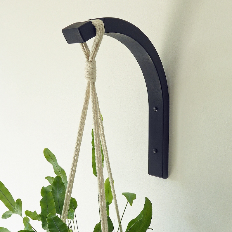 Puede incluir: Un colgador de plantas de metal negro con una cuerda de macram&eacute; blanca colgando de &eacute;l. El colgador est&aacute; montado en una pared blanca.