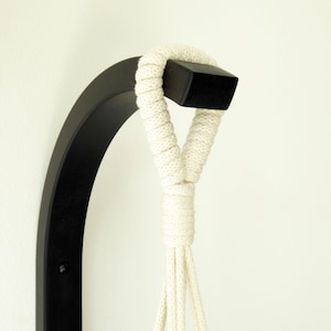 Puede incluir: Un colgador de plantas de pared de metal negro con una cuerda de algod&oacute;n blanca. La cuerda est&aacute; anudada en la parte superior del colgador.
