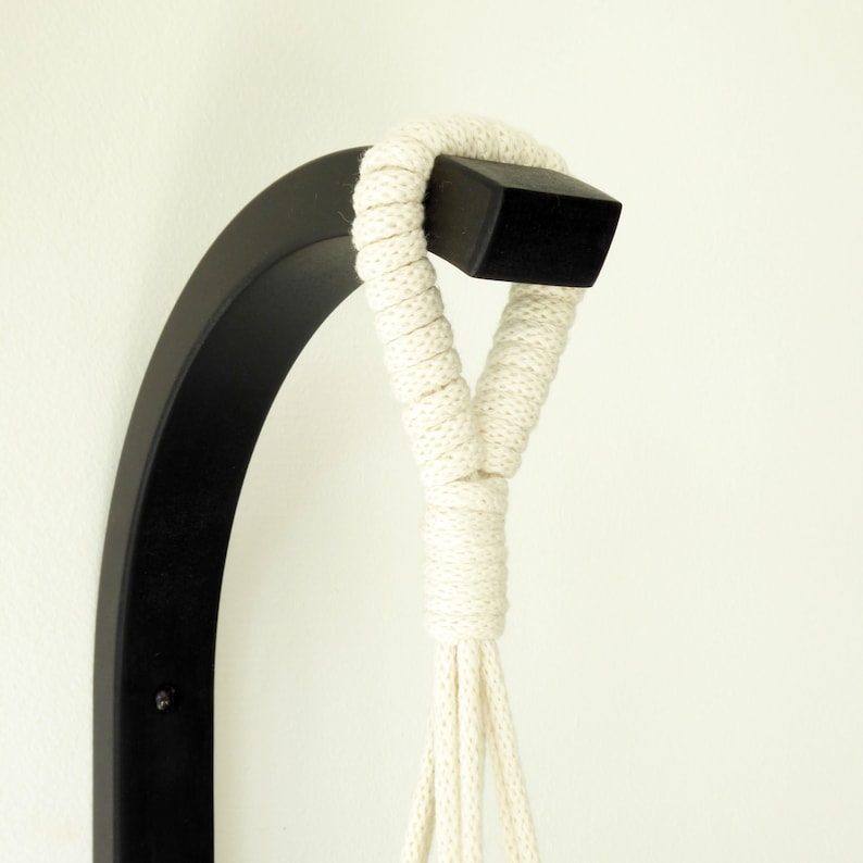 Puede incluir: Un colgador de plantas de pared de metal negro con una cuerda de algod&oacute;n blanca. La cuerda est&aacute; anudada en la parte superior del colgador.