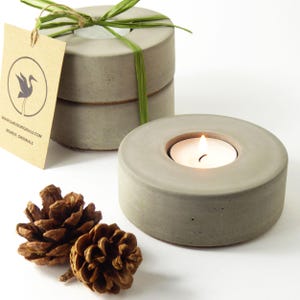 May include: A grey concrete candle holder with a lit tea light. Two more candle holders are stacked behind it, tied with green ribbon. A brown tag with a logo and website is visible. Two pine cones are in the foreground.