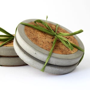 May include: A pair of round coasters with a grey concrete edge and a cork centre. They are tied with green raffia ribbon. The background is white. Each coaster measures approximately 10 cm in diameter.