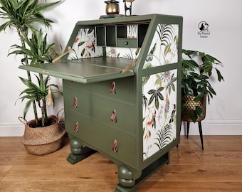 May include: A green vintage secretary desk with a drop-down front and three drawers. The desk is decorated with a white and green floral wallpaper featuring birds and leaves. The desk has leather pulls and sits on four turned legs. The desk is labeled "The Painted Toucan".