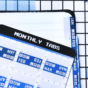 Könnte beinhalten: Ein Bogen mit selbstklebenden Monatsregistern mit weißem Text auf blauem Hintergrund, beschriftet von JAN bis JUN. Der Bogen trägt die Überschrift MONTHLY TABS in weißem Text auf schwarzem Grund. Ein schwarzer Stift liegt auf einem karierten Notizbuch.