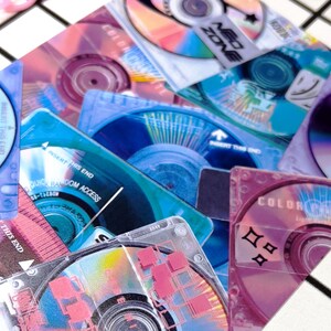May include: A collection of colorful mini CDs and cases in shades of blue, pink, and purple. Some cases have text like "NEO ZONE" and "COLOR PALETTE". The image has a retro, Y2K aesthetic.
