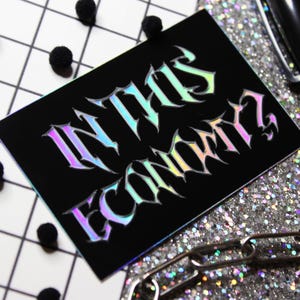 Puede incluir: Una pegatina negra con letras hologr&aacute;ficas iridiscentes que dicen "IN THIS ECONOMY?"