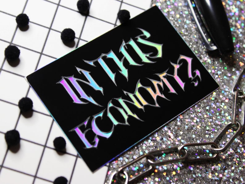 Puede incluir: Una pegatina negra con letras hologr&aacute;ficas iridiscentes que dicen "IN THIS ECONOMY?"