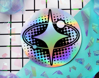 May include: A circular holographic sticker featuring a black graphic design of a four-pointed star centered within an oval ring and two small orbiting spheres, all set against a background of black halftone dots on a reflective surface.