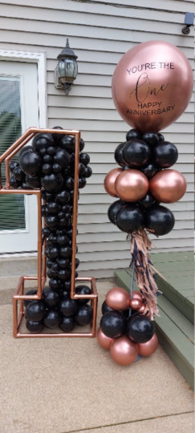 Puede incluir: Un gran globo de color oro rosa con el texto "You're the One Happy Anniversary" en escritura negra. Un arco de globos negro y oro rosa en forma del n&uacute;mero uno.