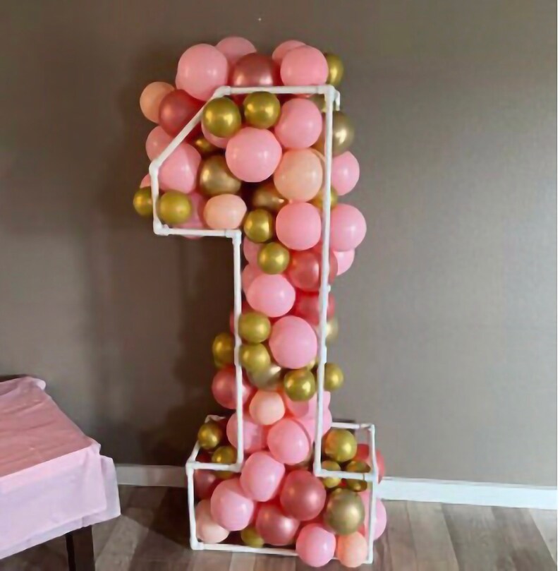 Puede incluir: Un gran n&uacute;mero uno hecho de globos. Los globos son rosas, dorados y melocot&oacute;n. El n&uacute;mero uno est&aacute; de pie sobre una superficie blanca.