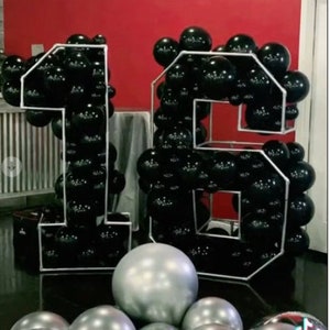 Puede incluir: Globos negros y plateados dispuestos en forma del n&uacute;mero 16. Los globos est&aacute;n en un marco blanco.