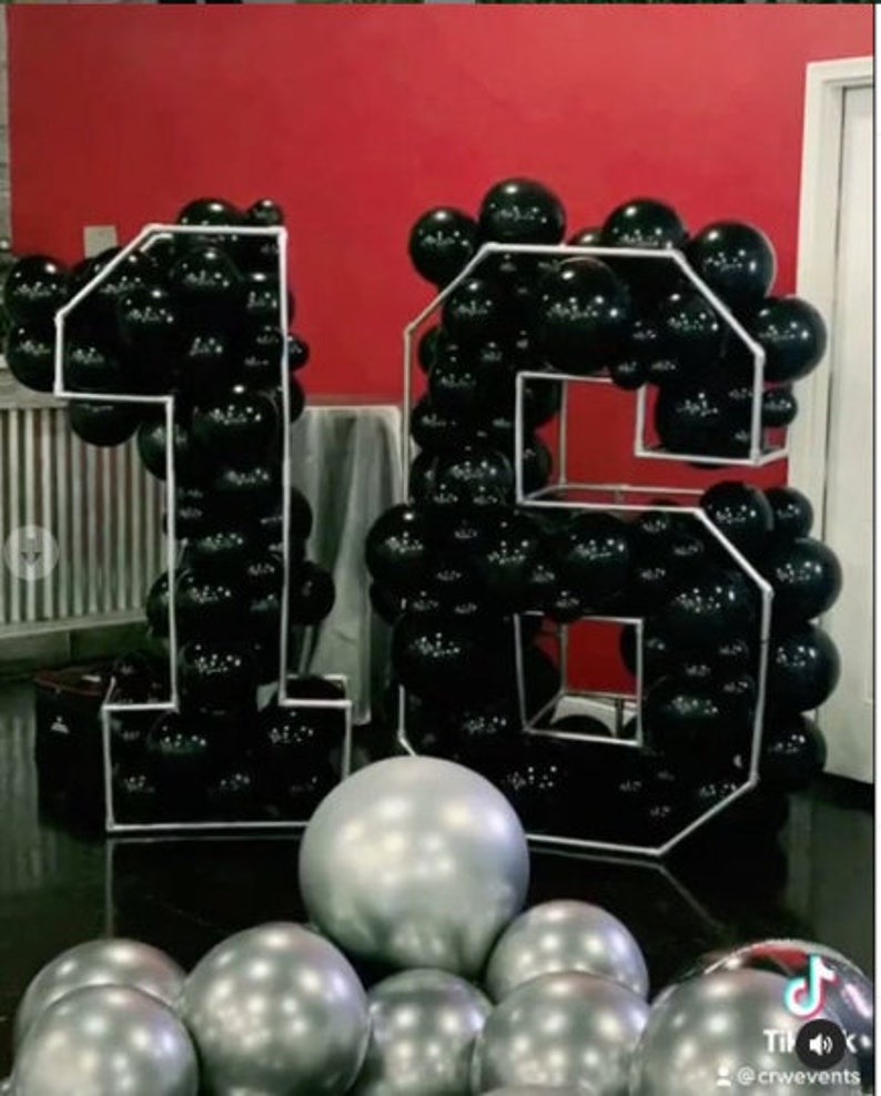 Puede incluir: Globos negros y plateados dispuestos en forma del n&uacute;mero 16. Los globos est&aacute;n en un marco blanco.