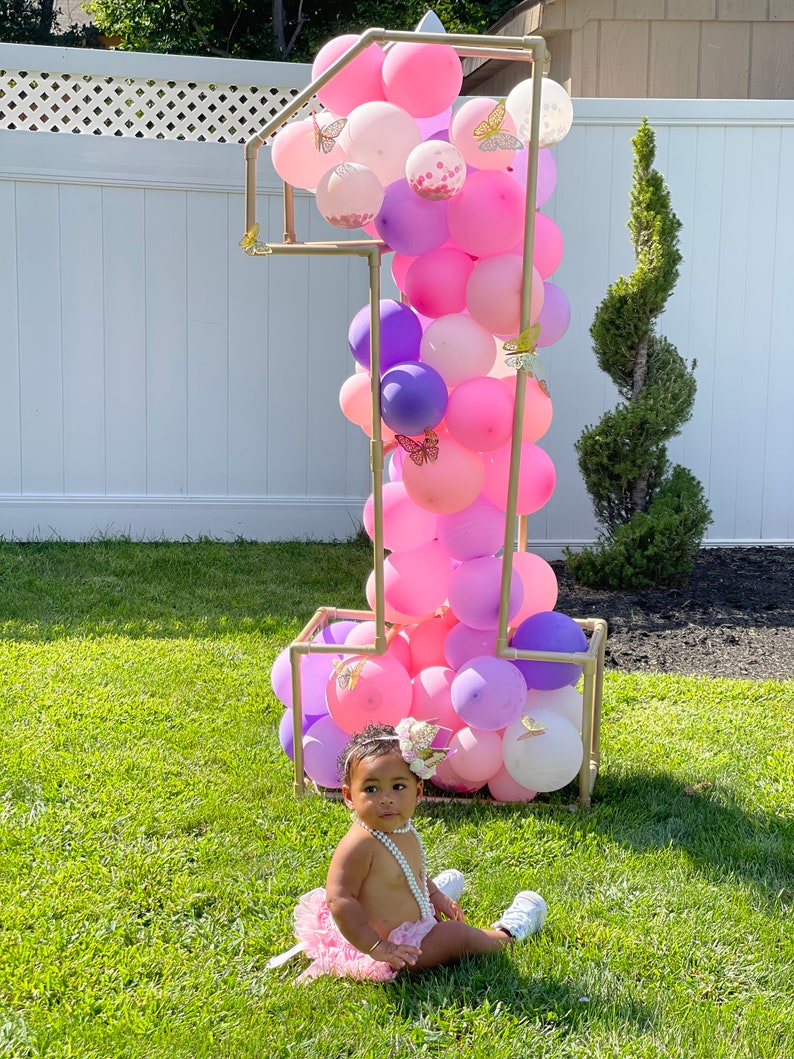 Puede incluir: Una ni&ntilde;a peque&ntilde;a con un tut&uacute; rosa y un collar de perlas se sienta frente a un gran n&uacute;mero uno hecho de globos. Los globos son rosas, morados y blancos con decoraciones de mariposas doradas. El n&uacute;mero uno est&aacute; hecho de tuber&iacute;as dor&eacute;es.